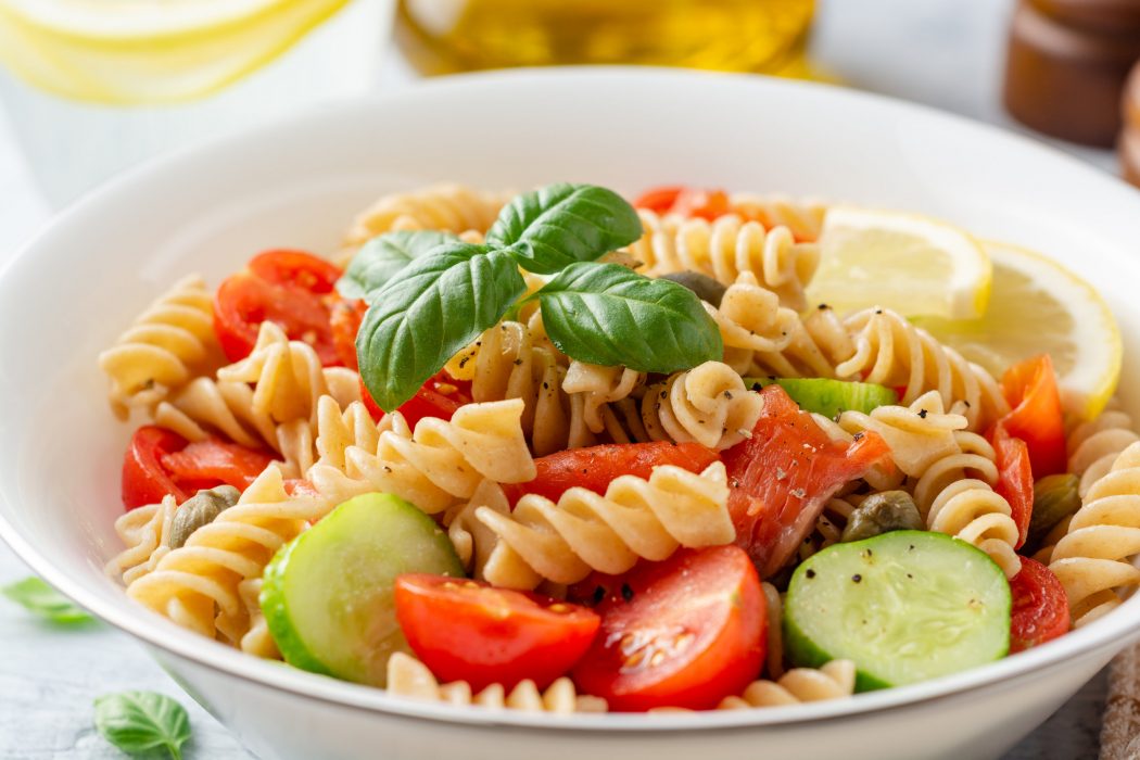 Whole wheat pasta salad with cucumbers, cherry tomatoes, salted salmon and capers on concrete background. Selective focus.