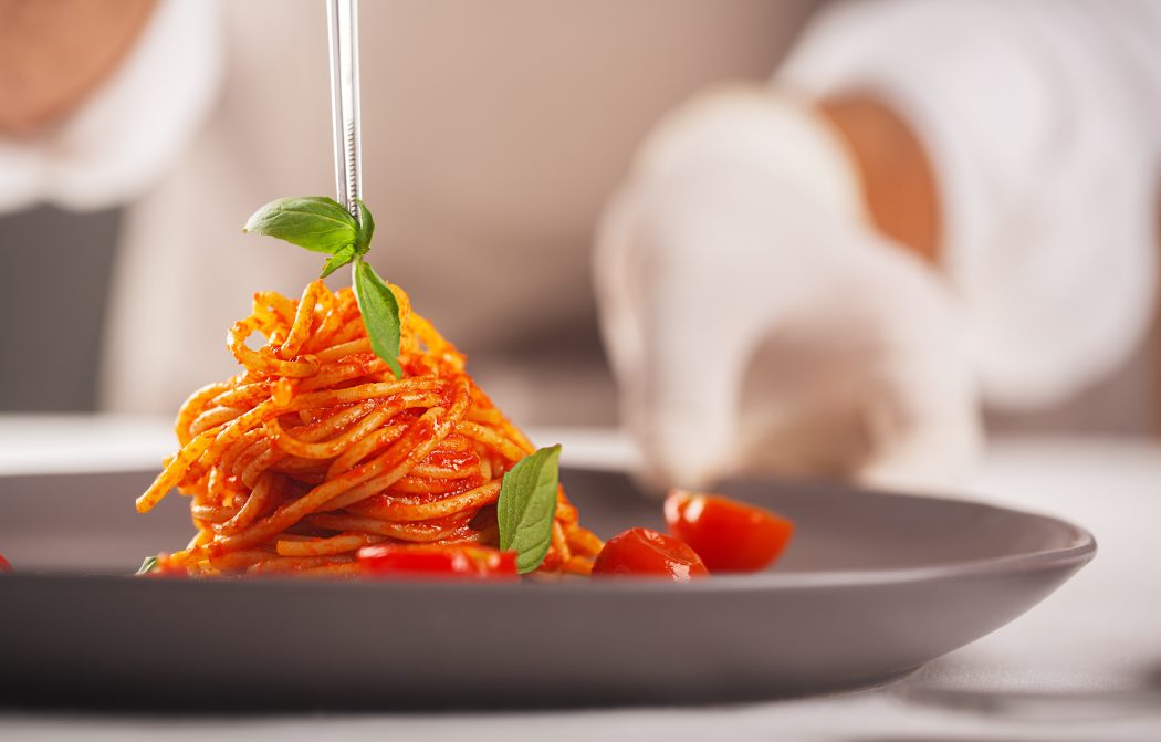 A chef in gloves and a white uniform is using tweezers to decorate a gourmet dish of pasta in tomato sauce with cherry tomatoes,plating food